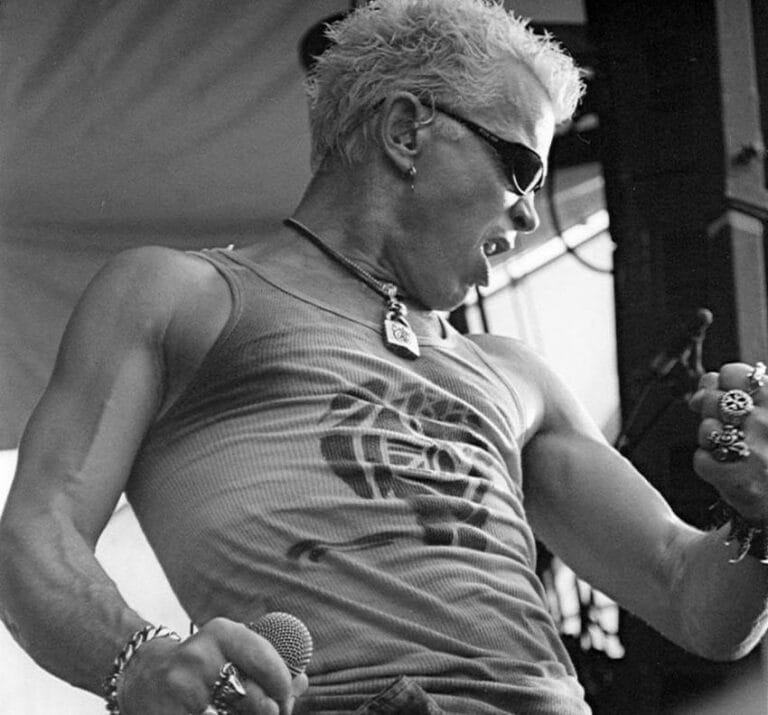 Billy Idol nastupa uživo na koncertu, prepoznatljiv po plavoj kosi i pank stilu